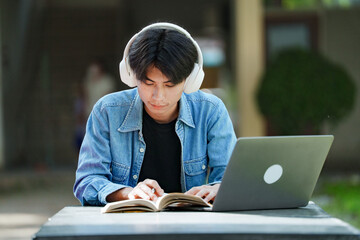 Portrait of a young Asian male college student e-learning using laptop on the bench on campus.