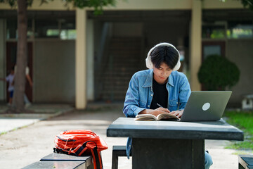 Portrait of a young Asian male college student e-learning using laptop on the bench on campus.