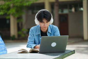 Portrait of a young Asian male college student e-learning using laptop on the bench on campus.