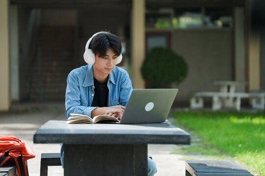Portrait of a young Asian male college student e-learning using laptop on the bench on campus.
