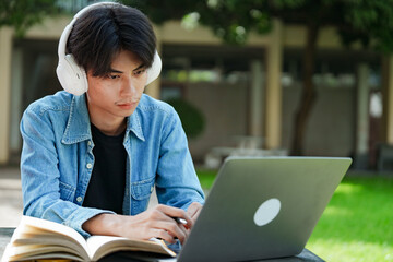 Portrait of a young Asian male college student e-learning using laptop on the bench on campus.