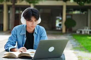 Portrait of a young Asian male college student e-learning using laptop on the bench on campus.