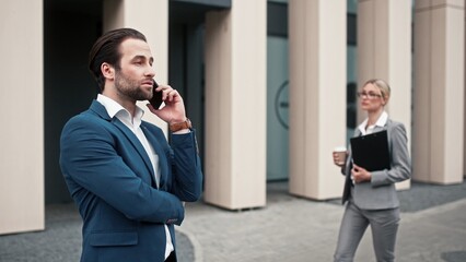 Male boss talking by phone outdoor. Female employee going past director. Businessman solving work issues. Office employees having break outside. Male in blue suit talking near office.
