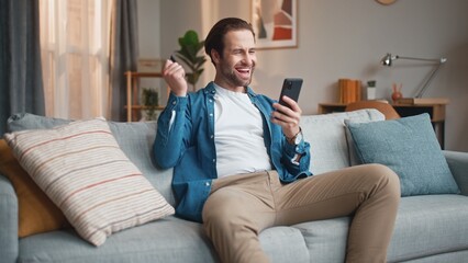 Obraz premium Cheerful man sitting on sofa and using smartphone. Male sitting on couch and shopping distantly. Happy man successfully finishing internet purchase. Male holding credit card and rejoicing.