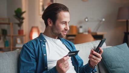 Fototapeta premium Man holding smartphone and cheerfully screaming. Male sitting on couch and shopping distantly. Happy man successfully finishing internet purchase. Male holding credit card and rejoicing.