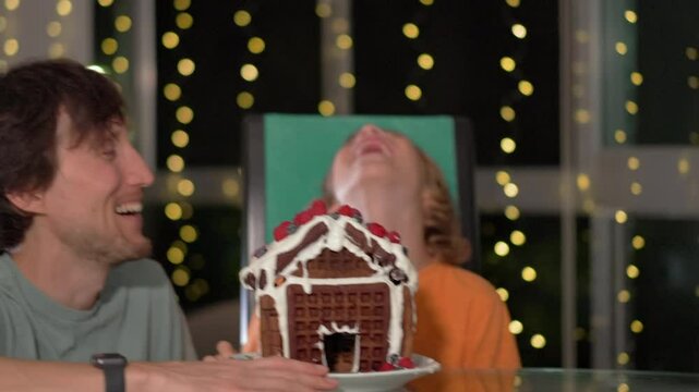 Joyful moments - father and son bite into the gingerbread house they decorated with fresh berries. Cozy winter evening, family fun, creativity, and festive bonding. Slowmotion video.