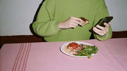 Person in green sweater using smartphone while eating sandwich with salad on pink tablecloth, minimalist aesthetic concept of digital distraction and modern solo mealtime habits
