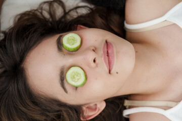 young caucasian female lies down with cucumber slices on both eyes enjoying refreshing skincare...