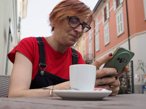 Youthful barista pausing during work shift, wearing red t shirt and dungarees, relaxing at outdoor cafe table while checking smartphone with blurred urban background