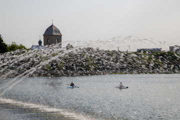 Athletes paddle kayaks on a calm lake with a city in the background. The water sparkles and the lake is surrounded by trees and a view of the city. The athletes compete against each other.