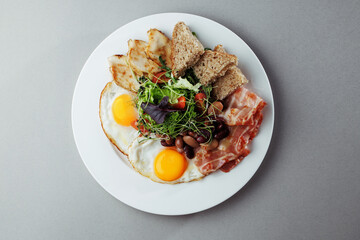 Healthy breakfast with eggs, bacon, grilled chicken, beans, salad and bread on white plate – top view gourmet morning meal on gray background
