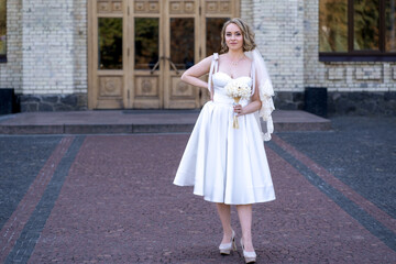 full length portrait of beautiful young bride in white short wedding dress and veil. Bride holding...