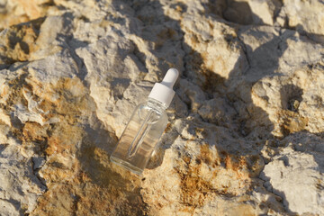 Glass dropper bottle with a clear serum placed on a textured rock surface near the sea. Natural...
