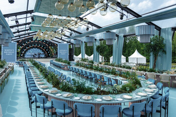 A lavish outdoor venue set for a birthday celebration, featuring long tables adorned with elegant plates and lush greenery. The expansive setting is framed by a transparent roof and decorative lightin