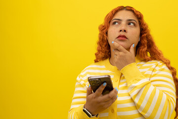 Freckled woman with curly red hair, thoughtful with hand on face while holding her cell phone on a yellow background with space for text