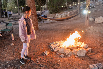 A young girl stands by a campfire in the forest at night while her family gathers nearby. A softly lit campsite with a hammock captures the warmth of a peaceful retreat.