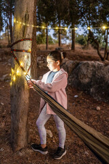 In a serene forest, a child sets up a hammock while string lights illuminate the area. Warm evening tones envelop the campsite, creating a peaceful atmosphere.