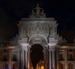 Night shot of the illuminated Arco da Rua Augusta in Lisbon, an 18th-century ornate triumphal arch with carved figures and views of the city centre.