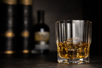 Whisky glass with ice cubes on dark surface, bottle and books in background