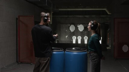 Medium long back view of male Caucasian instructor standing at distance from interactive bullseye, holding and aiming handgun while communicating with female trainee on indoor range