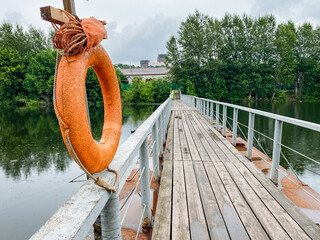 A bright orange lifebuoy sits on an old dock by calm waters, surrounded by lush greenery and colorful foliage, creating a peaceful atmosphere perfect for relaxation and adventure in nature