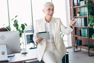 Elegantly dressed senior businesswoman with white short hair interacting at office desk holding a...