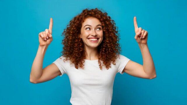 Excited Young Woman Pointing Upwards - A cheerful young woman with curly red hair points upwards with both index fingers against a bright blue background. She is wearing a white t-shirt and looks exci