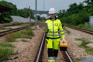 A man in a yellow and green safety suit is walking on railroad tracks. He is holding a laptop and a...