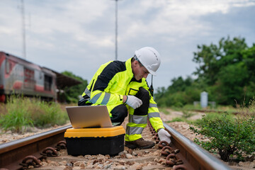 A man in a yellow and black safety suit is working on a train track. He is using a laptop and a...