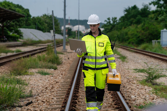 A man in a yellow and green safety suit is walking on railroad tracks. He is holding a laptop and a toolbox. Inspection and checking of railway tracks, railway maintenance.