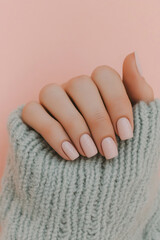 Subtle beauty: A feminine hand with delicately polished nails in a pale pink shade, contrasting softly with a cozy pastel knitted sweater. Ideal for concepts of wellness, natural beauty and self-care.
