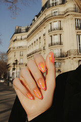 Stylish urban manicure: Hands with vibrant orange and pink gradient nails, highlighting a modern look in a sunny city environment. Ideal for beauty, fashion, travel and cosmopolitan lifestyle themes.
