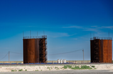 The photo shows two large rusty storage tanks standing in an open area under a clear blue sky. Between them are power poles and elements of industrial infrastructure.