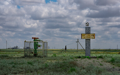 The photo shows a steppe landscape with an oil wellhead fenced with mesh and a memorial sign topped...