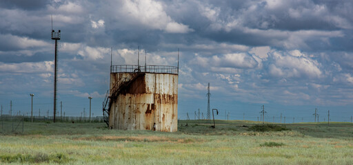 The photo shows a rusty cylindrical oil storage tank standing in an open field. Surrounding it are steppe, power lines, and a watchtower against a dramatic cloudy sky.