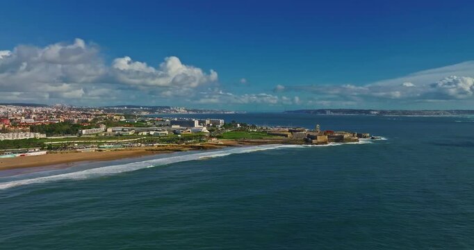Drone view of a fortress in Portugal with the famous bridge in the background. Saint Julian Fortress with lighthouse tower from beach of Carcavelos, Portugal. Atlantic ocean waves beach in Carcavelos