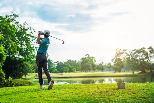 Confident Asian young man golfer swinging golf club powerfully on fairway under bright summer sunlight.