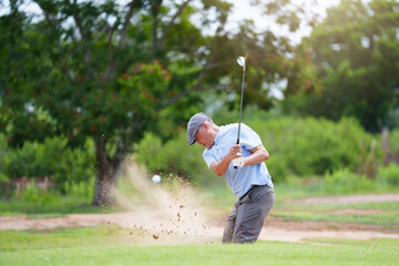 Asian Senior Man Golfer Confidently Swinging Golf Club on Sand Trap with Iron Driver under Sunshine in Summer Vibes.