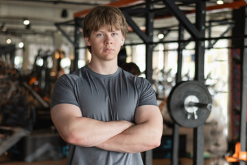 Handsome young personal trainer or fitness coach standing wearing gray t-shirt with cross hand at gym. Motivation for regular workout, body building, health and energy. Crafting my workout routine.