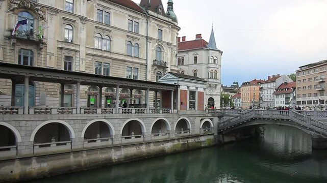 A vibrant daytime view of the charming Ljubljanica River flowing through the heart of Ljubljana, Slovenia. The Triple Bridge (Tromostovje), a unique and iconic landmark designed by Jože Plečnik