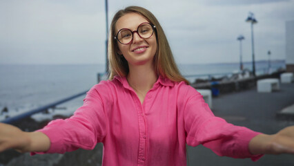 Woman in pink shirt smiling by the sea on a beach promenade with arms open wide, capturing joy and...