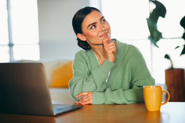 Charming young woman in a cozy green cardigan smiling thoughtfully while enjoying leisure time at home in bright lighting