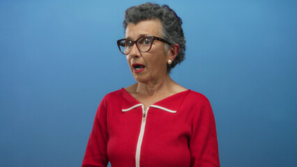 Senior woman with grey hair wearing red surprised against blue background, expressing shock and emotion, glasses accentuating her reaction.