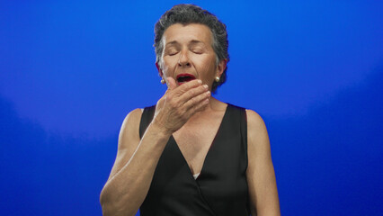 Senior woman with grey hair sneezing and covering her mouth against a vivid blue background,...