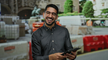 Smiling young man holding tablet at construction site with urban background and colorful barriers, embodying professional outdoor setting.