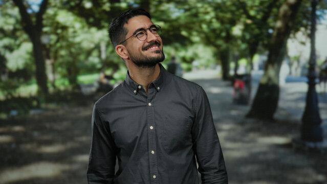 Young man with glasses in outdoor park setting, looking contemplative and stylish with green trees in the background, capturing a serene and thoughtful ambiance.