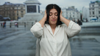 Woman in white shirt stands in vatican city square covering ears with a calm expression, portraying...