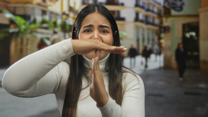 Young hispanic woman gesturing stop on an urban street surrounded by blurred pedestrians and...