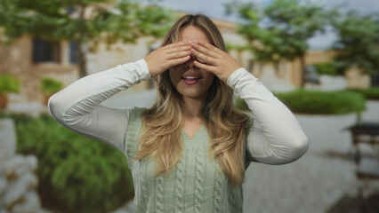 Woman covers her eyes with both hands in a lush green park setting under natural sunlight; surprise...