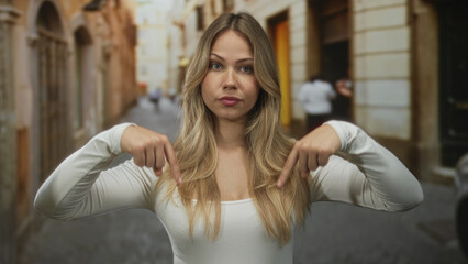Woman with long blonde hair pointing both index fingers down on a narrow cobblestone street; attention focus emphasis.